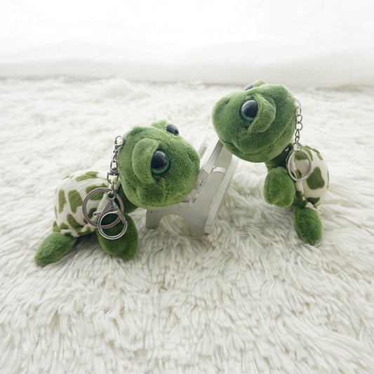Two green plush sea turtle keychains with patterned shells, resting on a white shag rug.