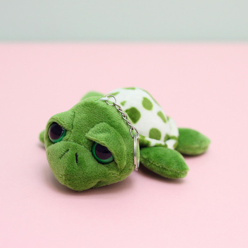 A small plush turtle keychain with a sad, downcast expression. The turtle is green with a white shell patterned with green spots, and has a silver metal keyring attached.