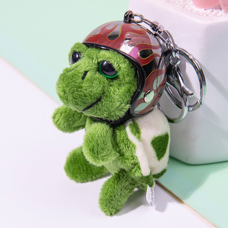 A plush green turtle keychain wearing a shiny red and black flame-patterned helmet with silver metal key rings, photographed against a pastel mint and white background.
